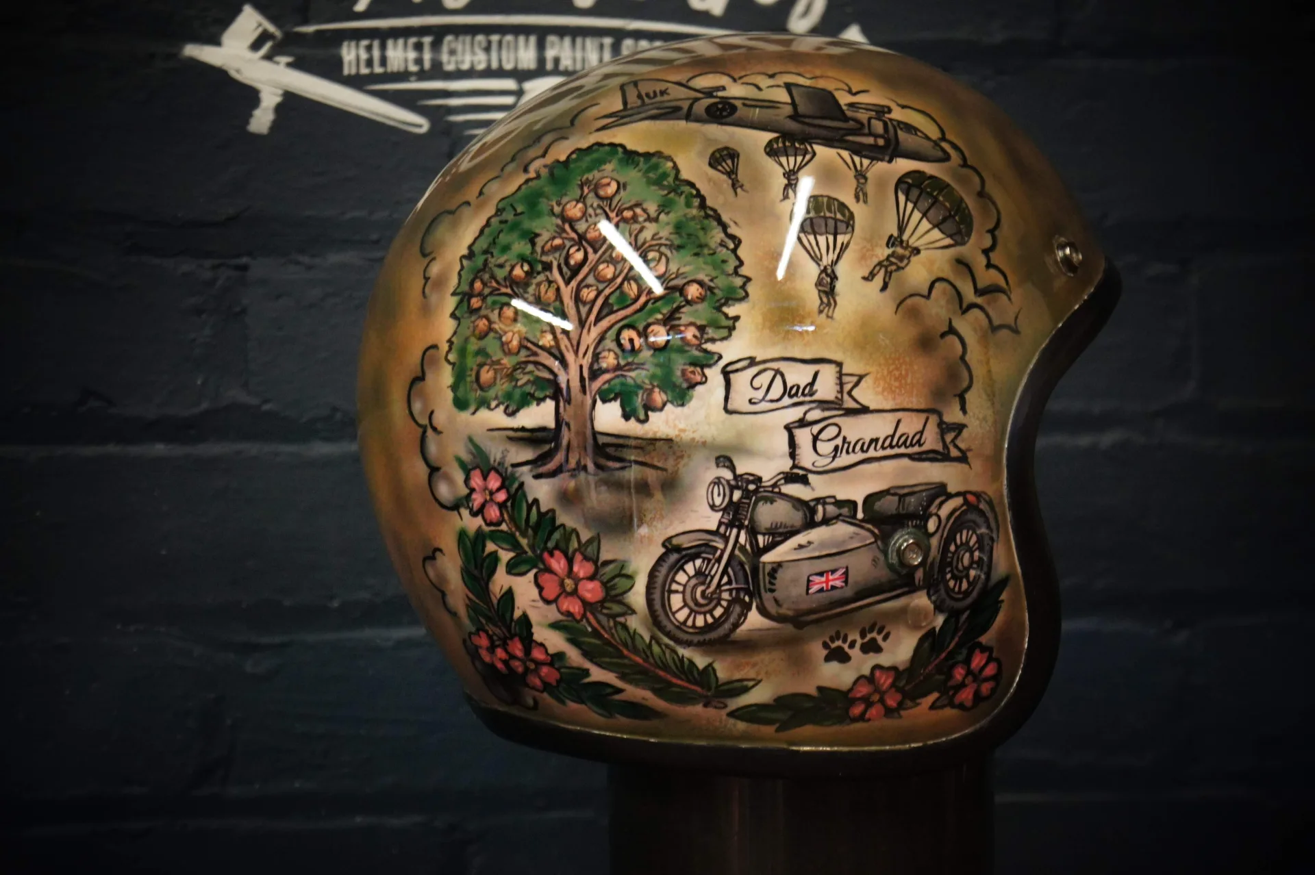 Left side of the helmet with the acorn-laden oak family tree, the 'Dad' and 'Grandad' banners, a motorcycle and sidecar, and paratroopers descending from a transport aircraft
