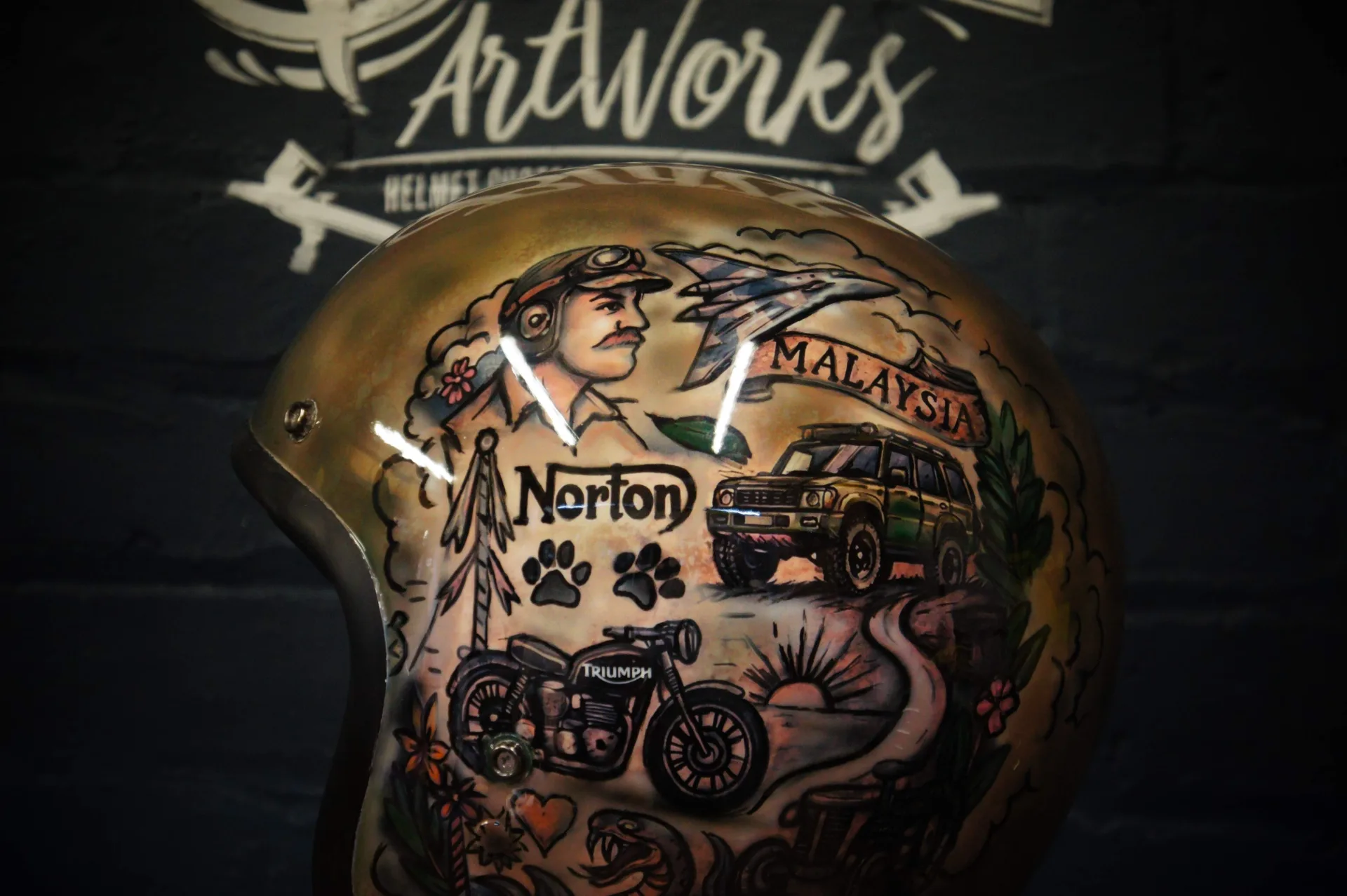 Front-left of the helmet featuring the aviator-capped portrait, a fighter jet, Norton and Triumph motorcycles, paw prints and a traditional-style snake