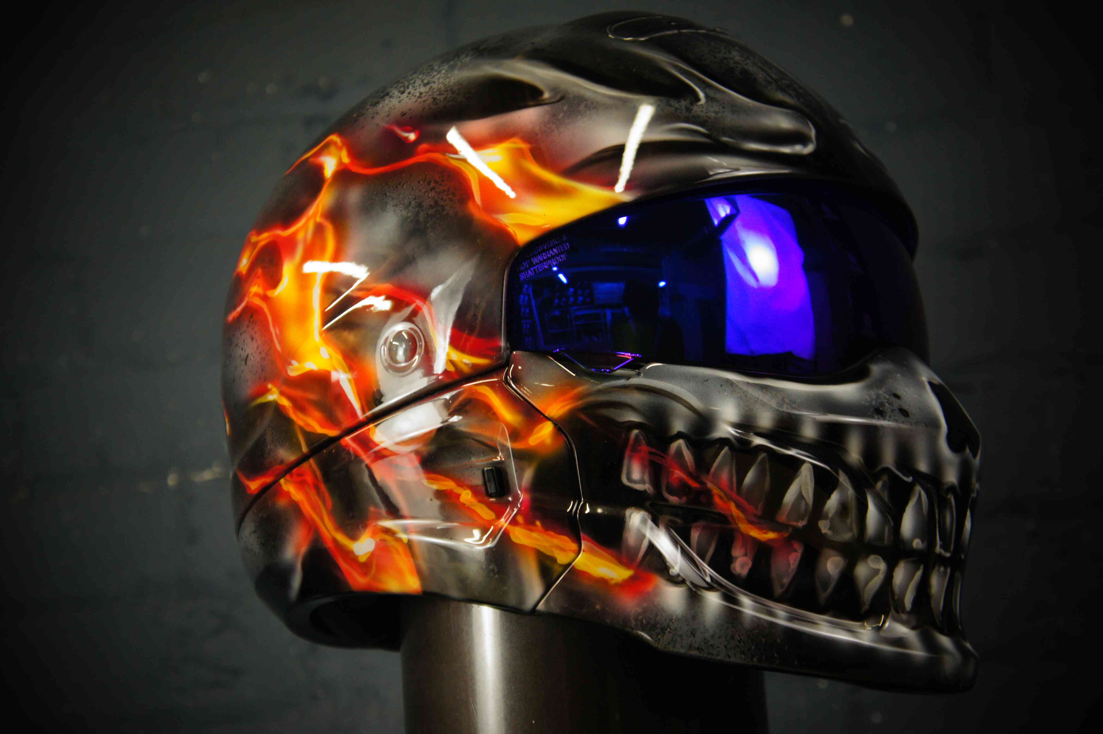 Black motorcycle helmet with orange fire flames surrounding metallic silver skull design, blue tinted visor, professional airbrush