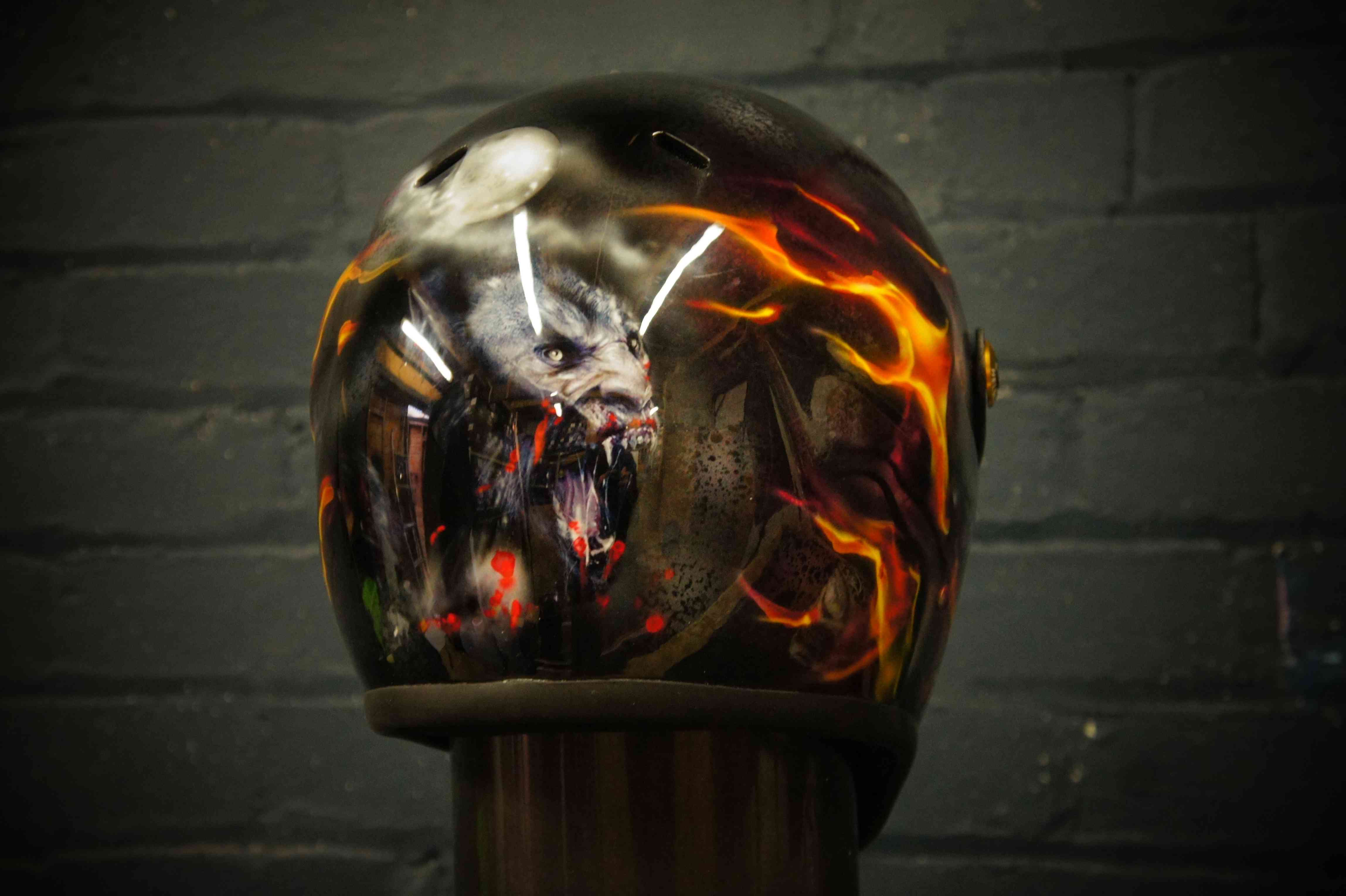 Black motorcycle helmet with demonic wolf creature surrounded by orange flames and red blood effects, professional airbrush art