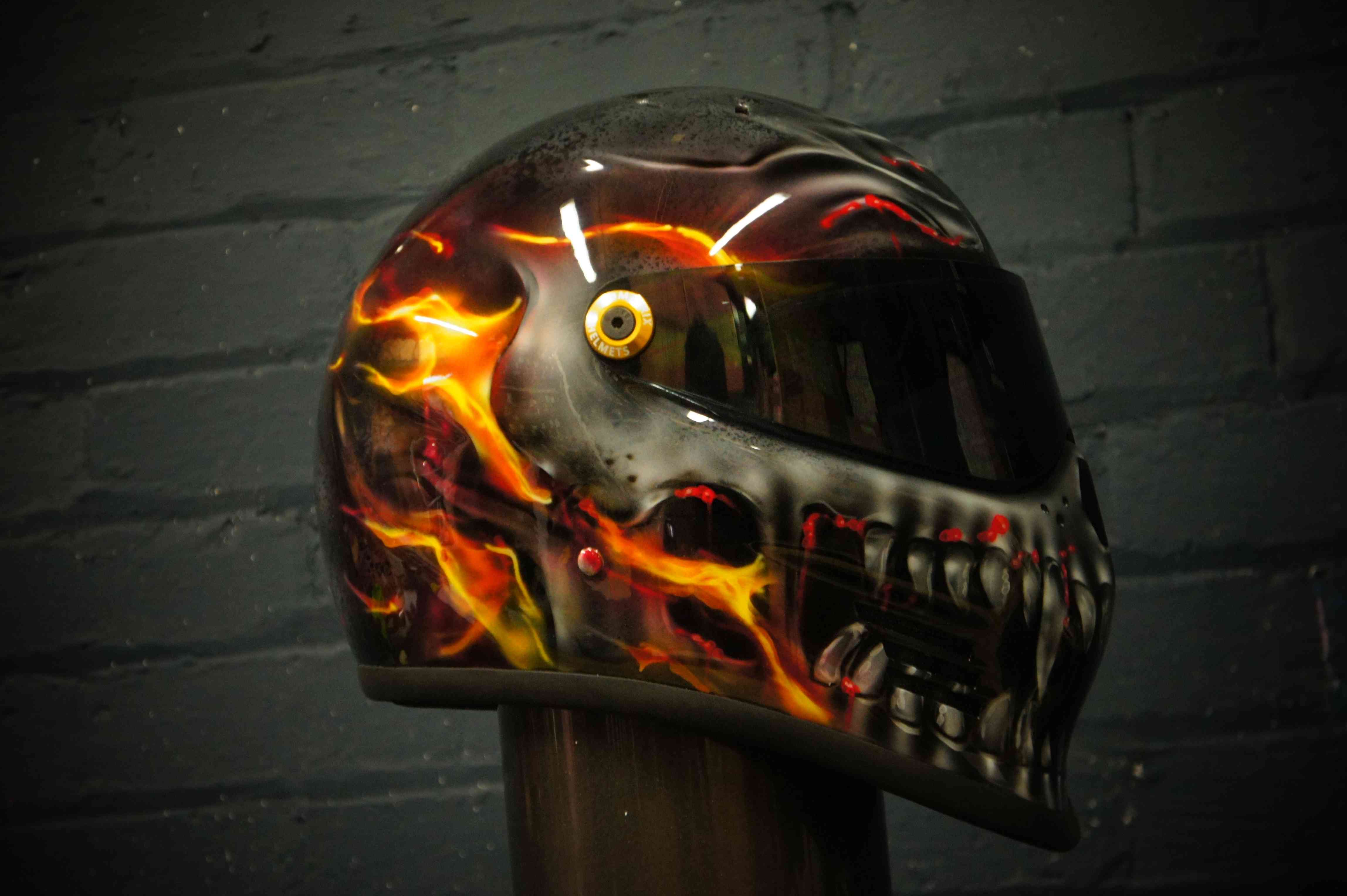 Black motorcycle helmet with airbrushed flaming skull design featuring orange and yellow fire effects, glowing eye detail
