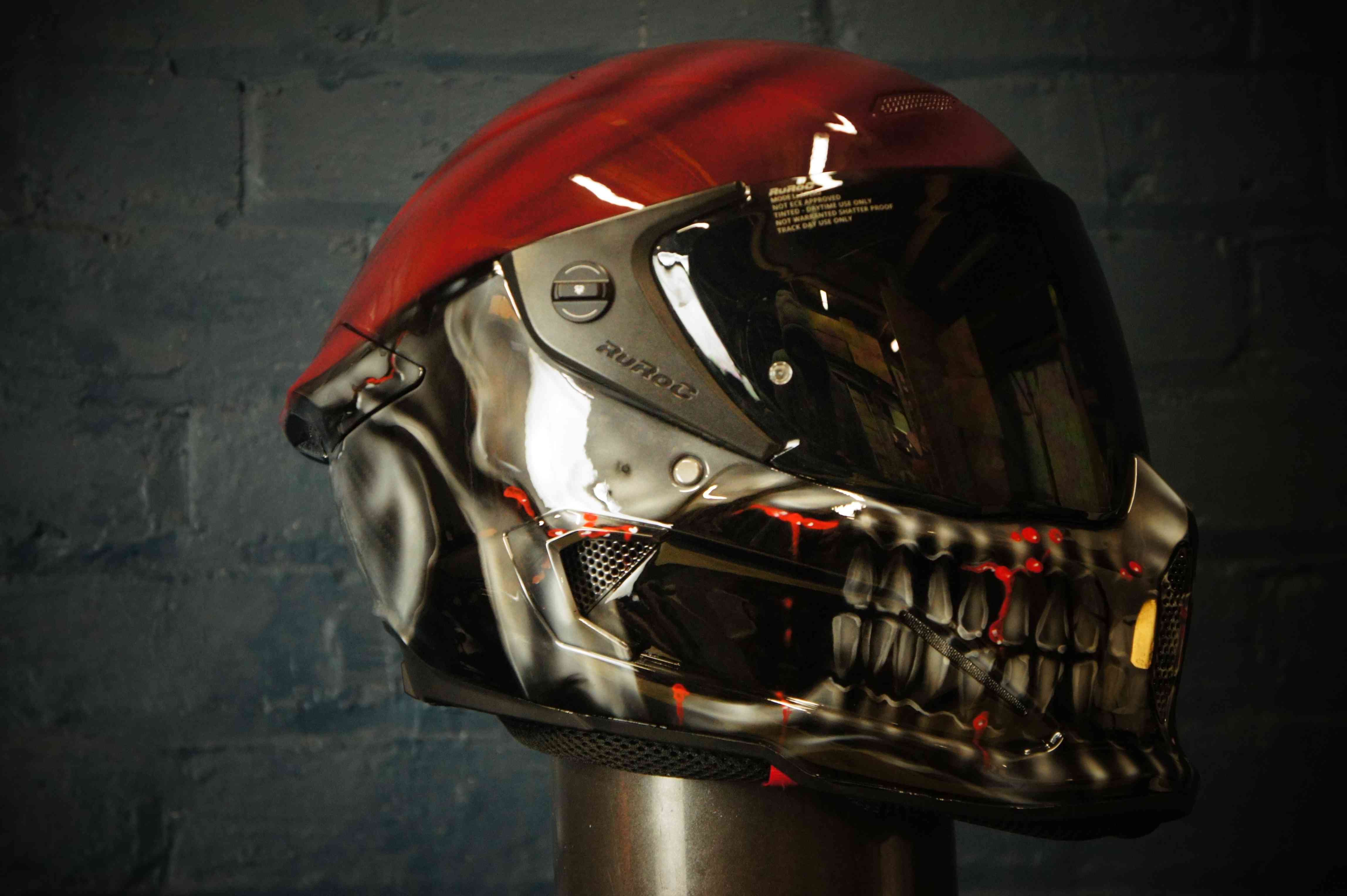 Black motorcycle helmet with photorealistic skull face design featuring glowing red eye sockets and detailed bone structure