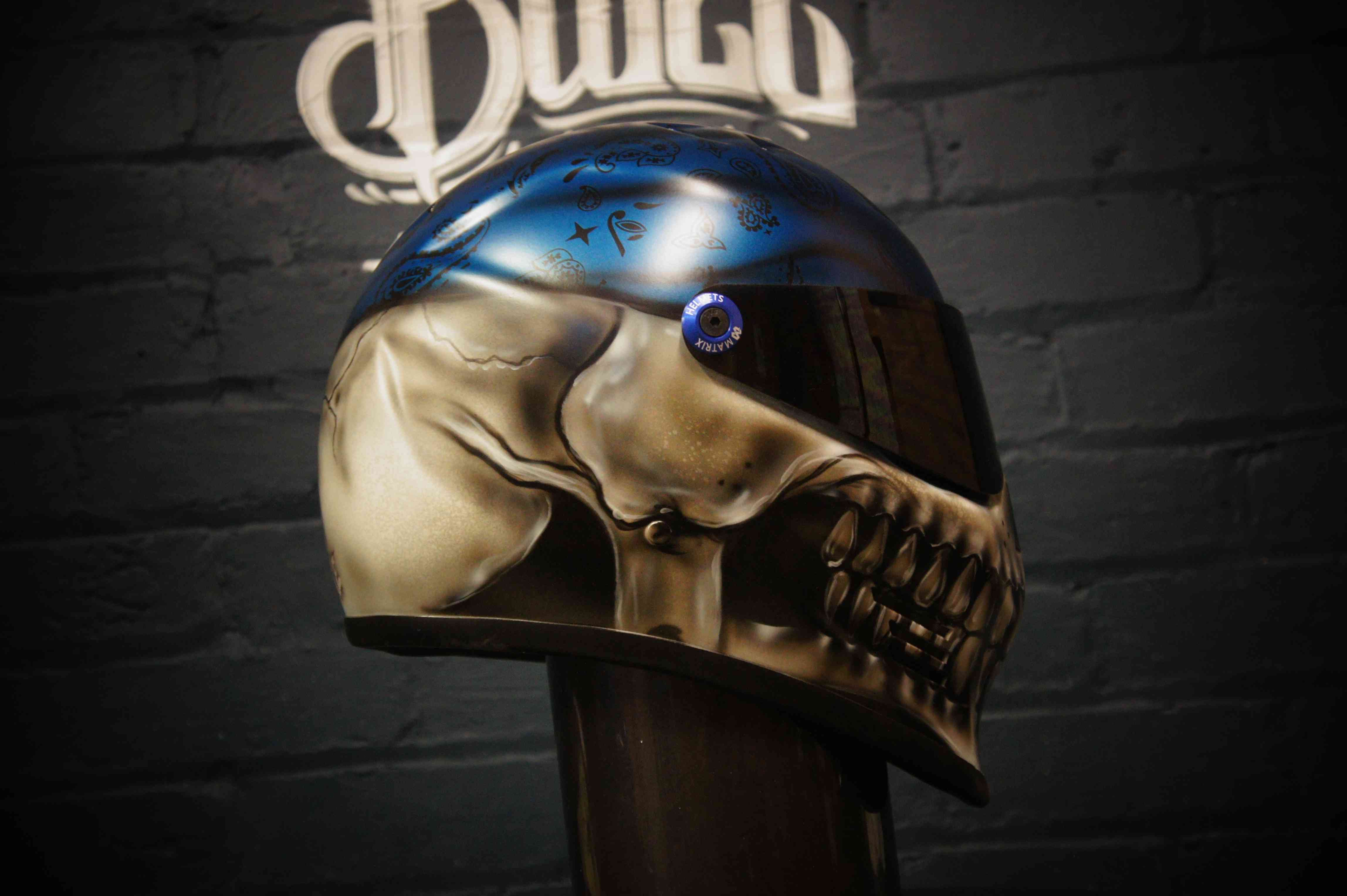 Photorealistic skull motorcycle helmet with metallic gold skull face, blue bandana with paisley pattern, glowing blue eye