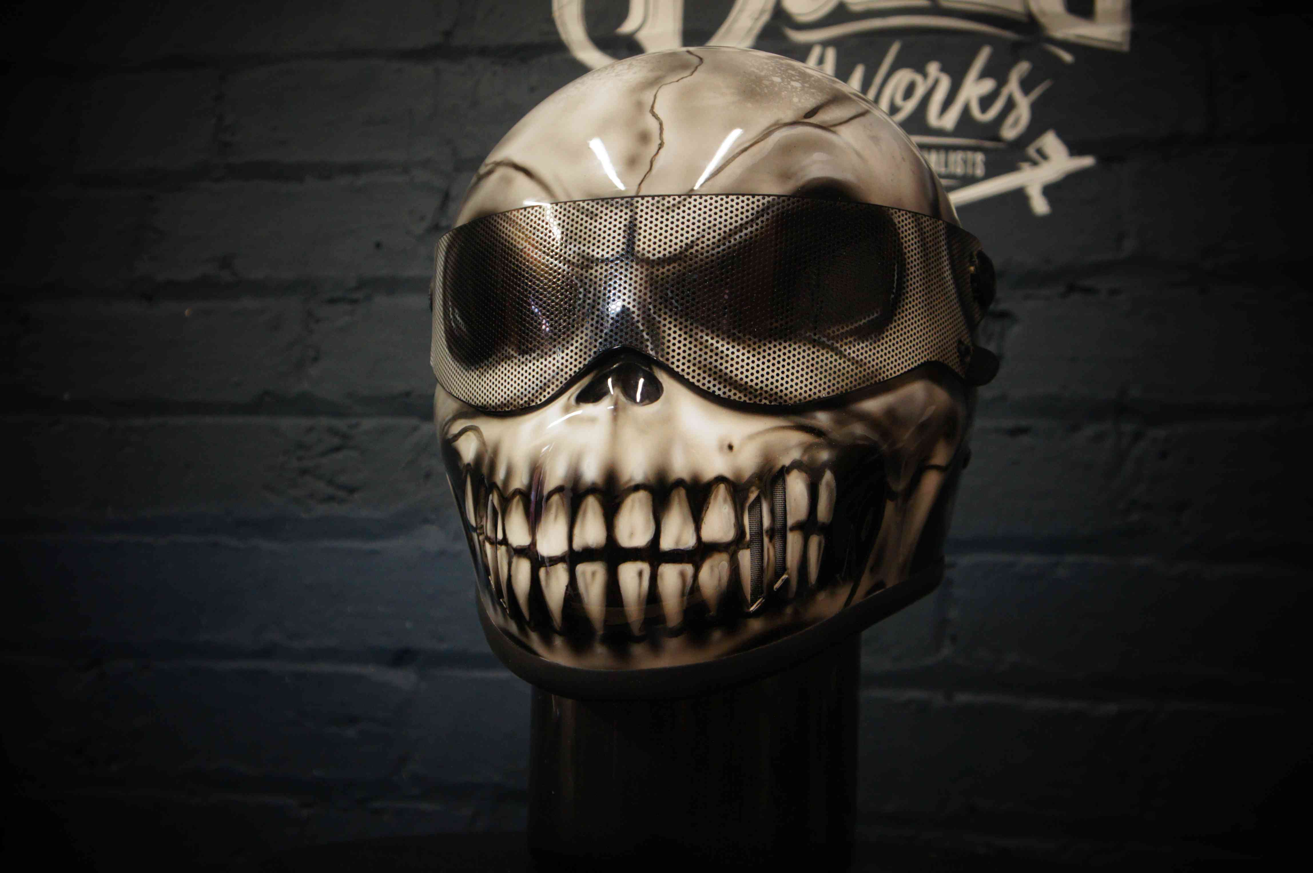 Dark grey motorcycle helmet with detailed grinning skull design featuring prominent white teeth, mesh goggles, and bone structure