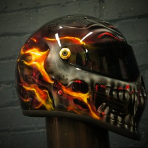 Black motorcycle helmet with airbrushed flaming skull design featuring orange and yellow fire effects, glowing eye detail