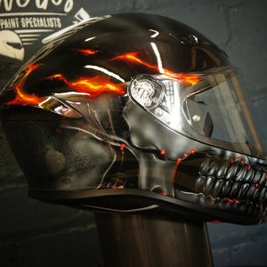 Black motorcycle helmet with realistic orange and red flames flowing over detailed skull design, featuring skeletal jaw