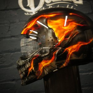 Black motorcycle helmet with realistic orange and red flames surrounding detailed chrome skull artwork in photorealistic style