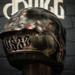 Black motorcycle helmet with gothic skull design featuring cracked stone texture, red glowing cracks and ornate lettering