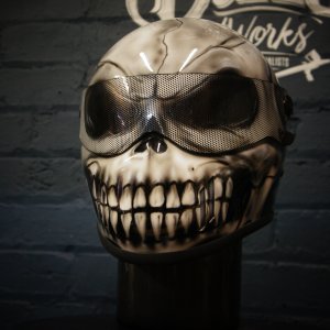 Dark grey motorcycle helmet with detailed grinning skull design featuring prominent white teeth, mesh goggles, and bone structure